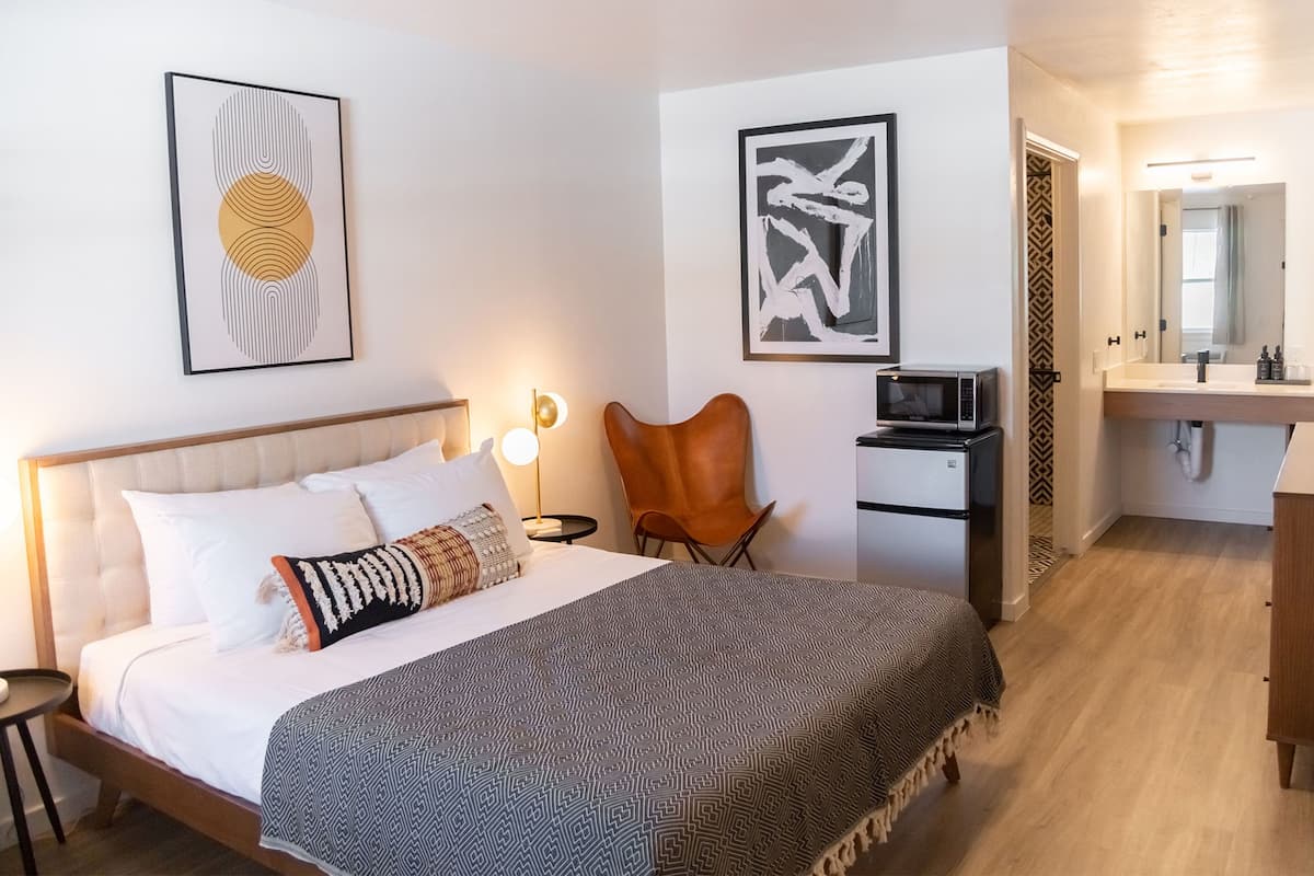A modern hotel room with a bed, a chair, a desk with a microwave, and a bathroom visible through an open door.