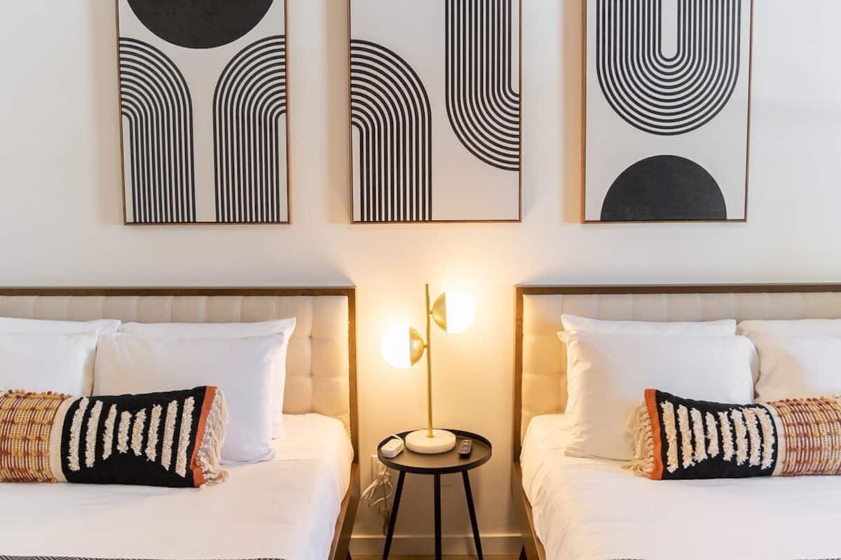 A hotel room with two beds, each with white linens and decorative pillows. There are three framed abstract artworks on the wall above the beds.