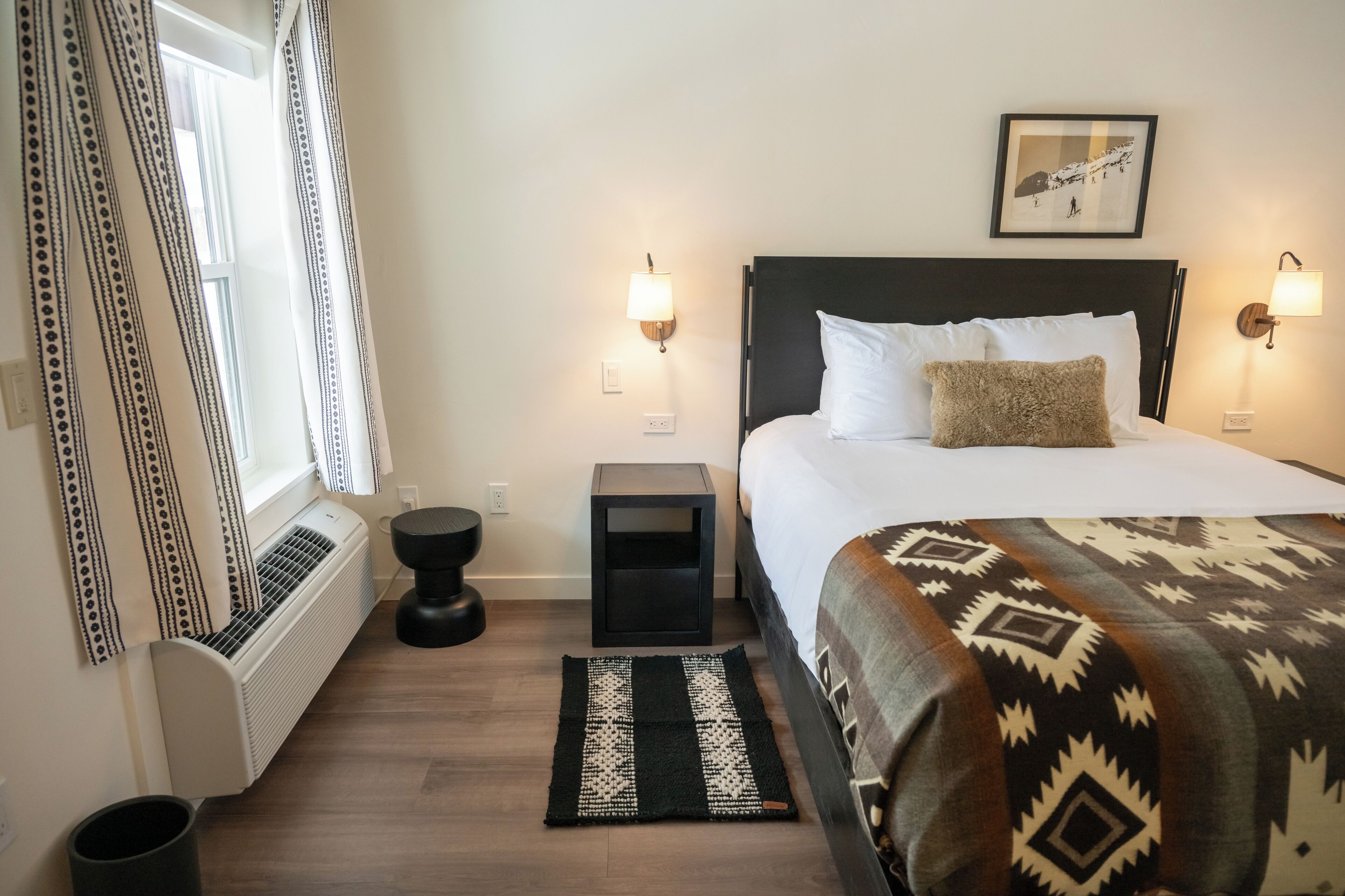 Une chambre d’hôtel avec un lit, des lampes de chevet, une fenêtre avec des rideaux, un radiateur, une petite table et un tapis.