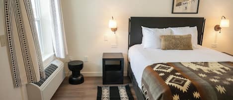 Hotelkamer met een bed, nachtlampjes, een raam met gordijnen, een radiator, een klein tafeltje en een vloerkleed.