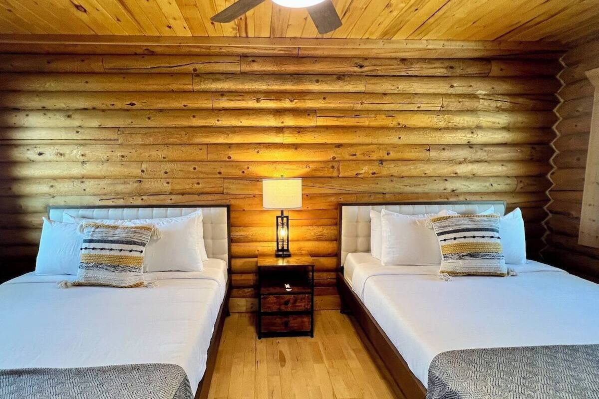 A log cabin bedroom with two beds, a wooden ceiling, and a nightstand with a lamp.