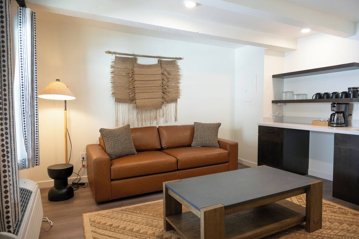 A modern living room with a brown leather sofa, a wooden coffee table, and a kitchen area with dark cabinets and a coffee machine.