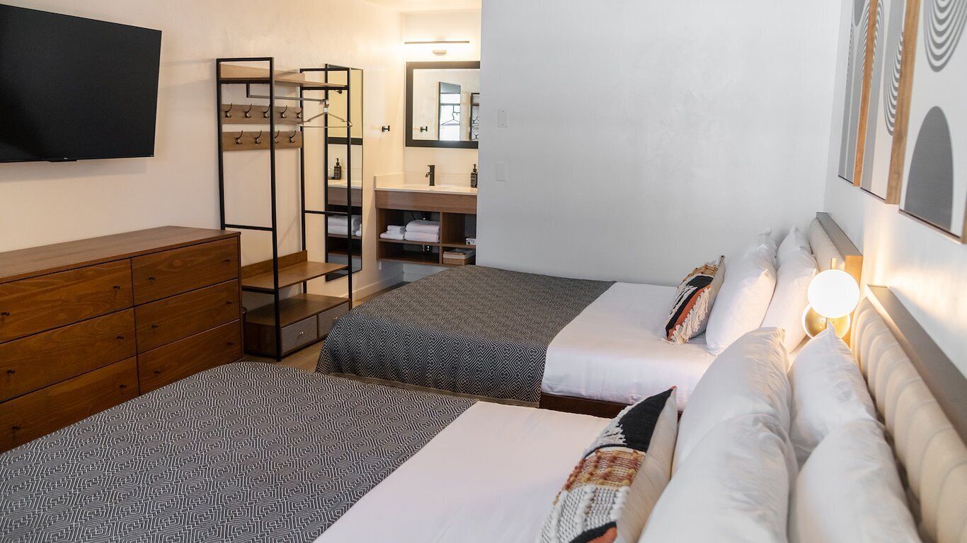 A hotel room with two beds, a wooden dresser, a TV, a mirror, and a bathroom with a sink and towel rack.