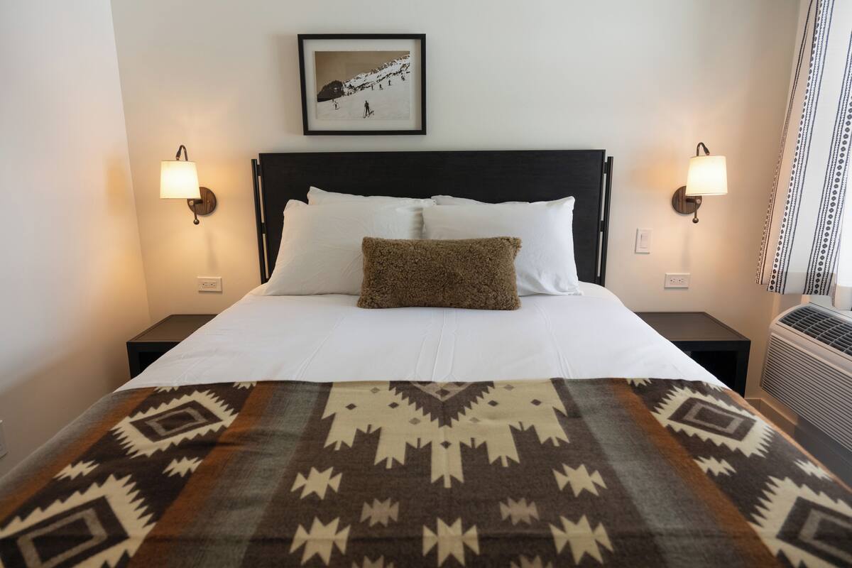 A hotel room with a large bed, two bedside lamps, a framed picture on the wall, and a patterned throw blanket.