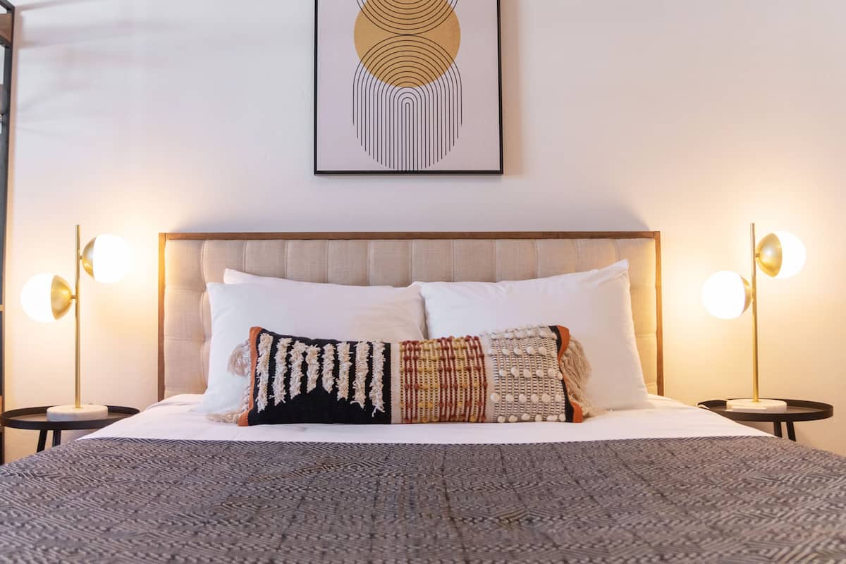 A neatly made bed with a patterned blanket, two bedside lamps, and a framed abstract artwork above the headboard.
