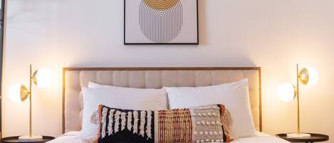 A neatly made bed with a patterned blanket, two bedside lamps, and a framed abstract artwork above the headboard.