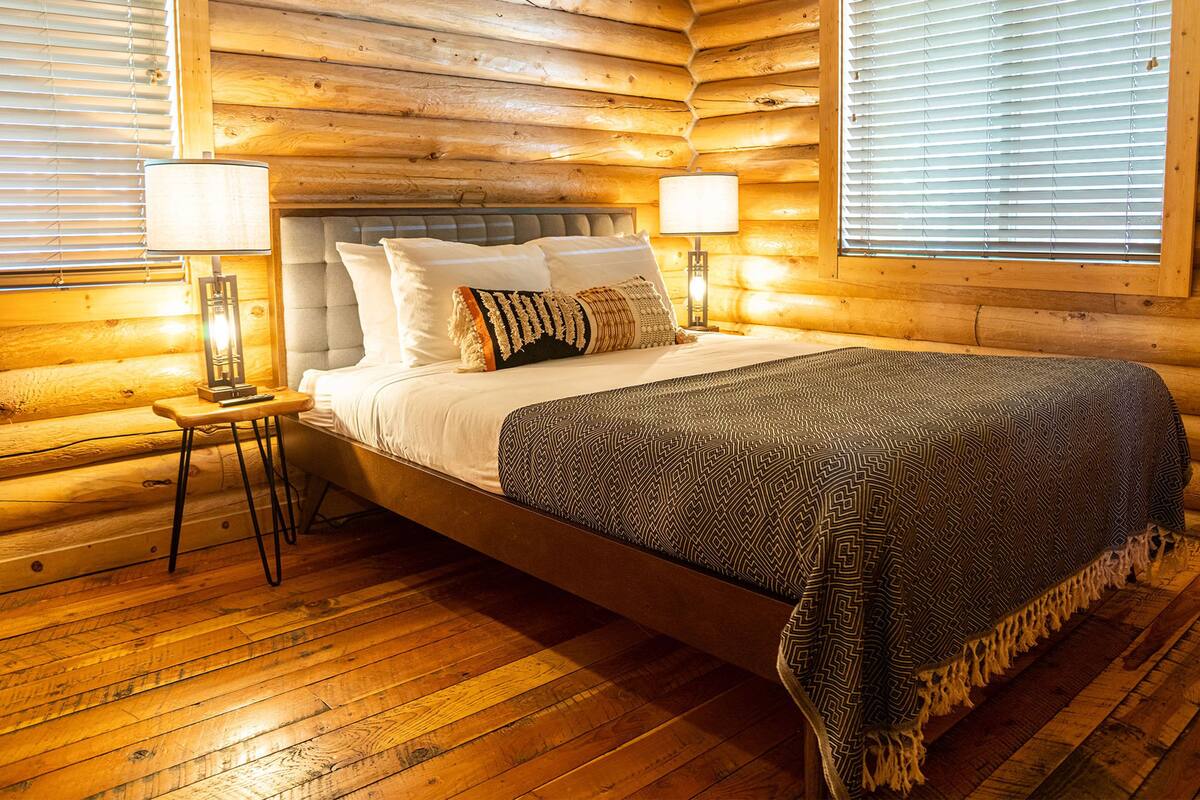 A log cabin bedroom with a bed, two bedside lamps, and a window with blinds.