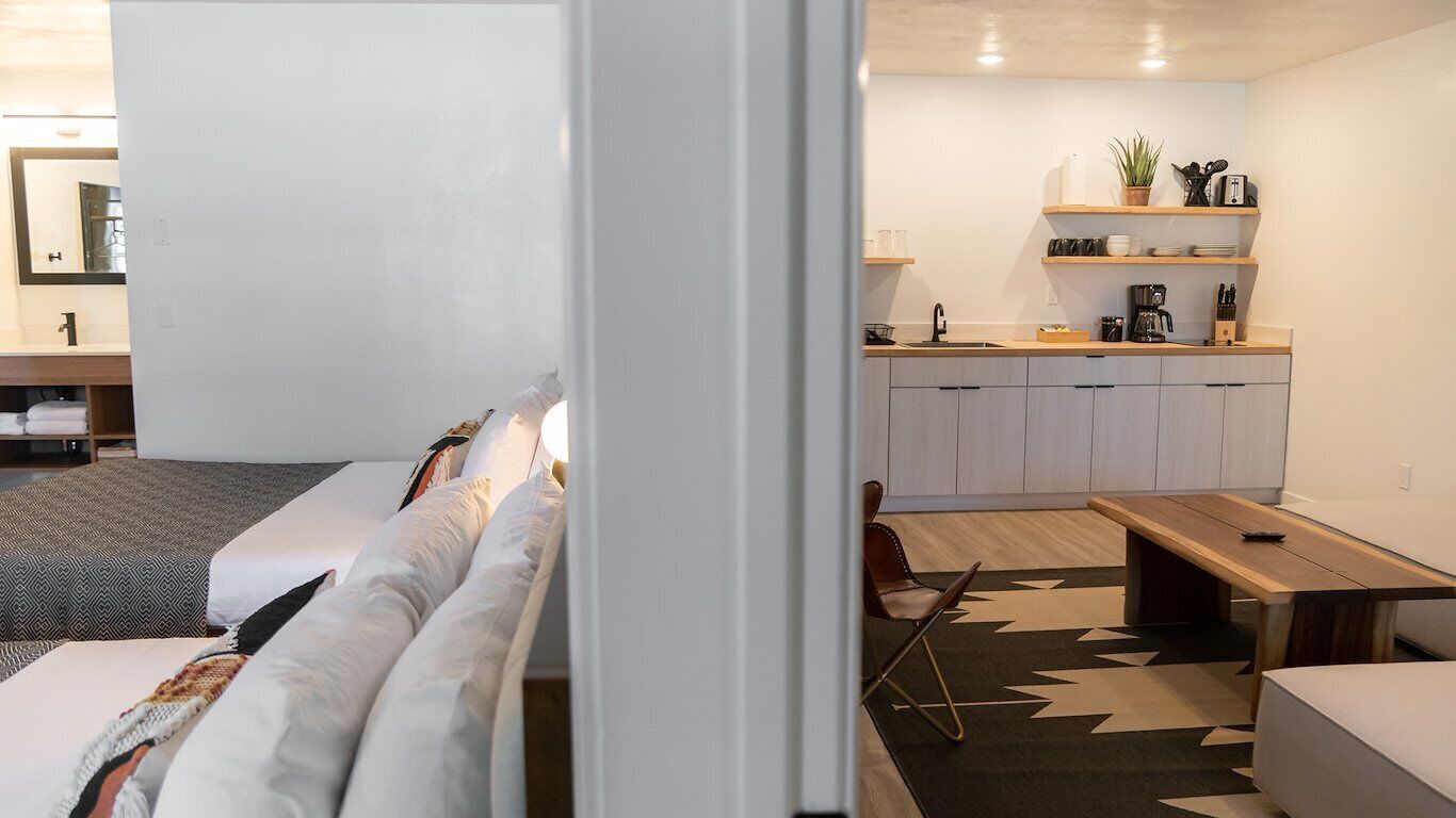 A modern living room with a sofa, a wooden coffee table, and a rug. Adjacent is a kitchen with white cabinets, a countertop, and a hanging plant.