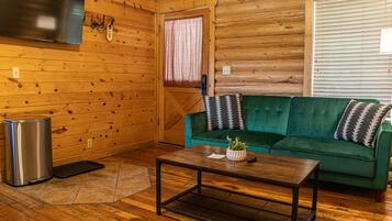 Un acogedor interior de cabaña de madera con un sofá verde, una mesa de centro de madera, un televisor montado en la pared y una ventana con persianas.