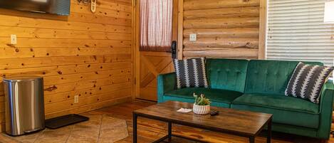 L’intérieur d’un chalet confortable, avec un canapé vert, une table basse en bois, un téléviseur fixé au mur et une fenêtre équipée de stores.