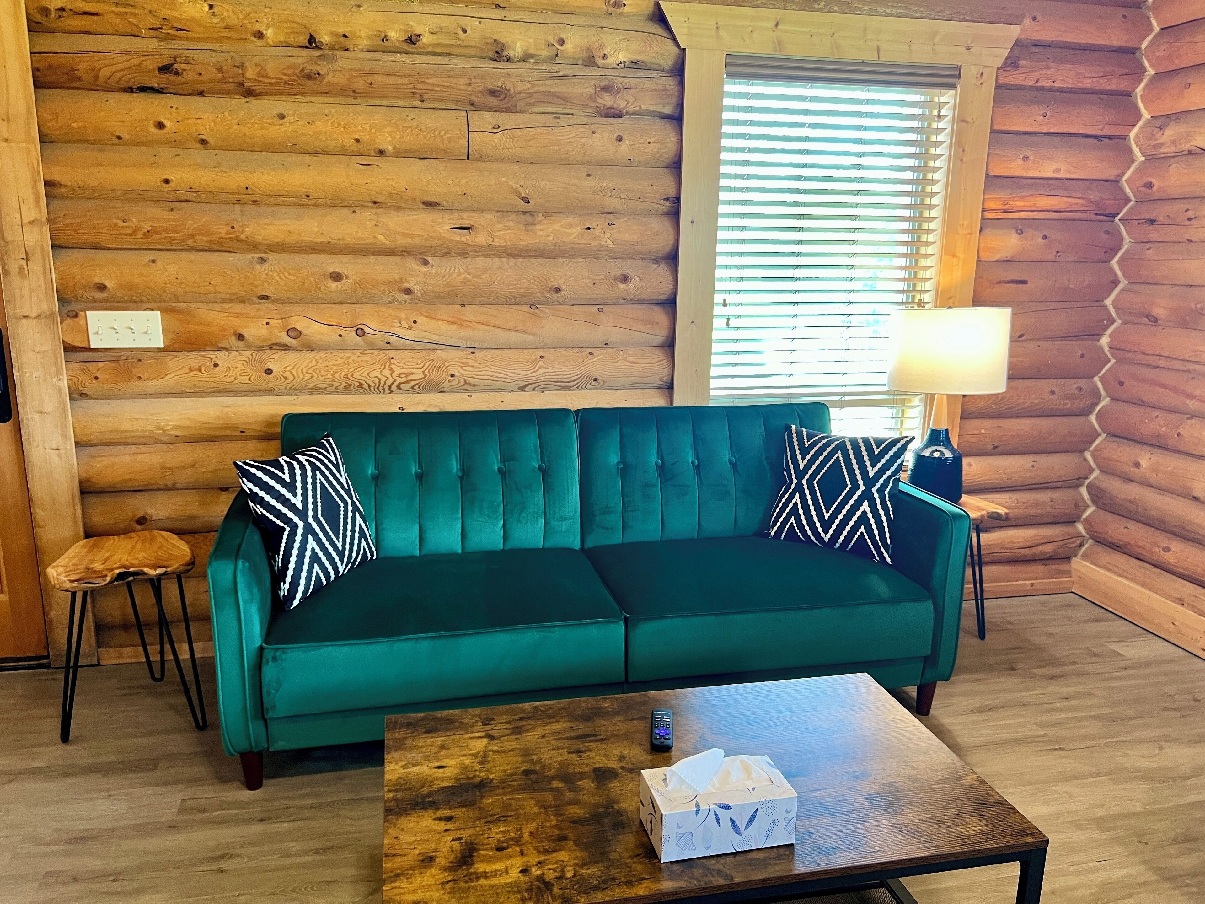 A log cabin interior with a teal sofa, two patterned pillows, a small wooden stool, a wooden coffee table with a tissue box, and a lamp on a side table.