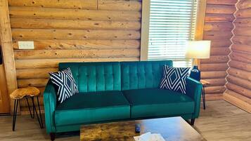 Interieur van een blokhut met een blauwgroene bank, twee gekleurde kussens, een klein houten krukje, een houten salontafel met een zakdoekendoos en een lamp op een bijzettafeltje.