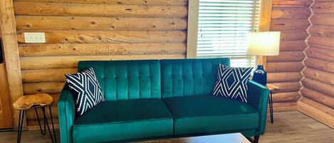 A log cabin interior with a teal sofa, two patterned pillows, a small wooden stool, a wooden coffee table with a tissue box, and a lamp on a side table.
