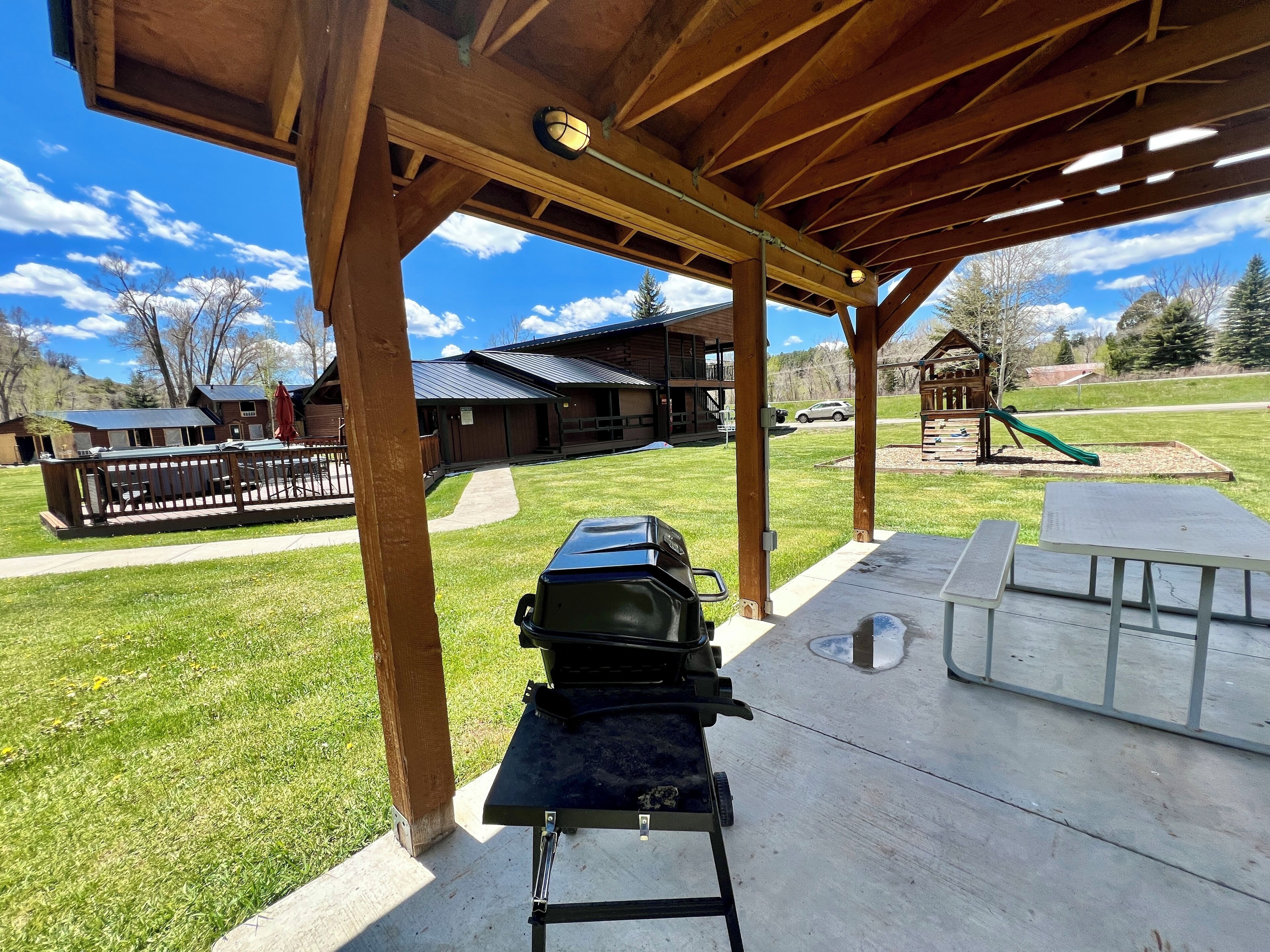 Ein überdachter Patio-Bereich mit Grill und Picknicktischen, im Hintergrund ein Spielplatz und ein Gebäude mit Metalldach.