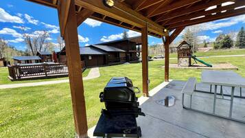 Ein überdachter Patio-Bereich mit Grill und Picknicktischen, im Hintergrund ein Spielplatz und ein Gebäude mit Metalldach.