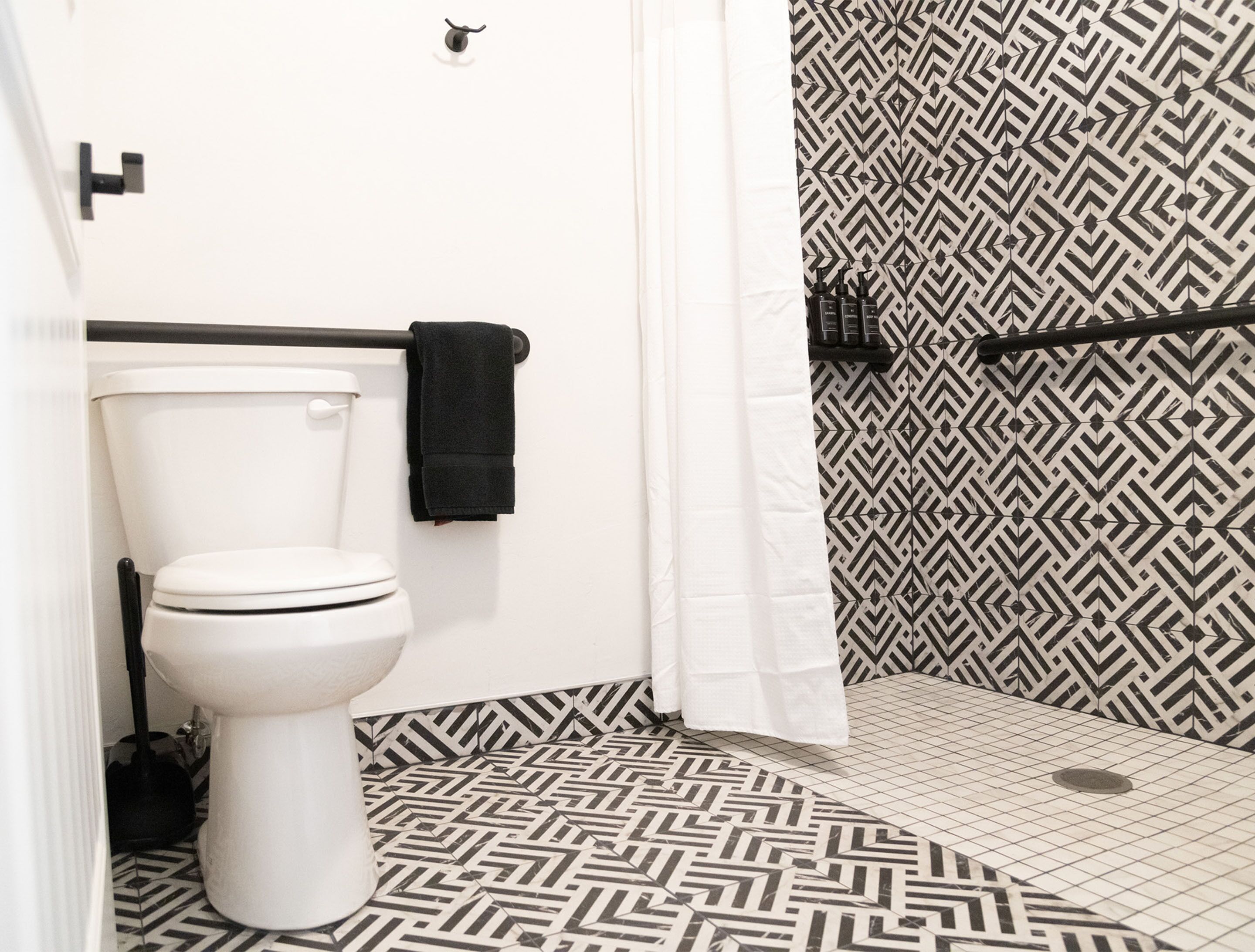 A bathroom with a white toilet, a black towel hanging on a rack, and a shower area with a black and white patterned shower curtain.