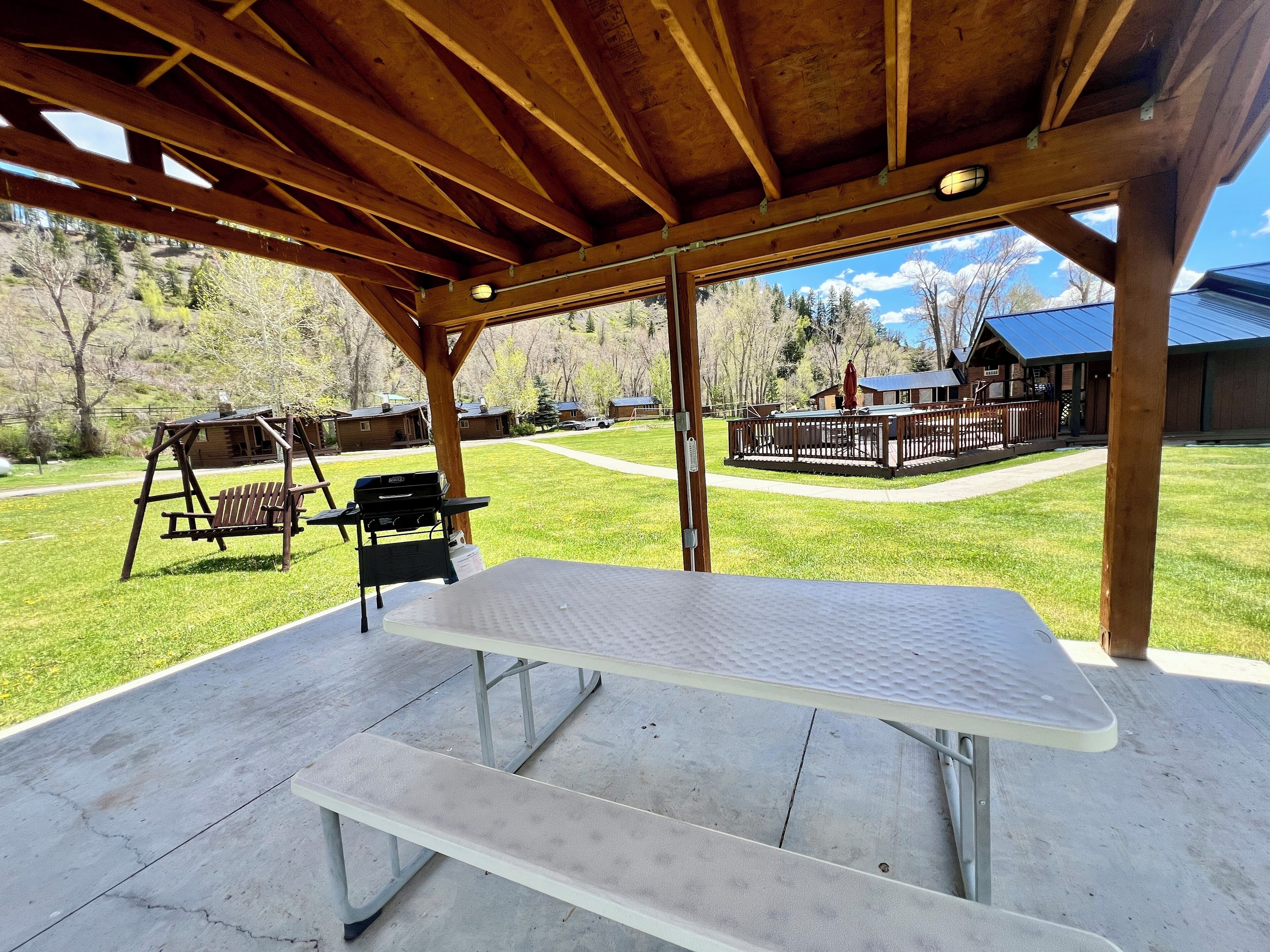 A covered picnic area with a table and benches, overlooking a grassy field and cabins.