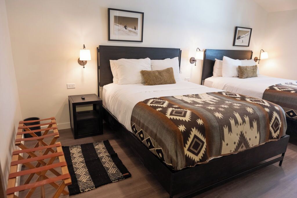 Habitación de hotel con dos camas, cada una con un edredón estampado, lámparas de noche, una mesita pequeña y un banco de madera.