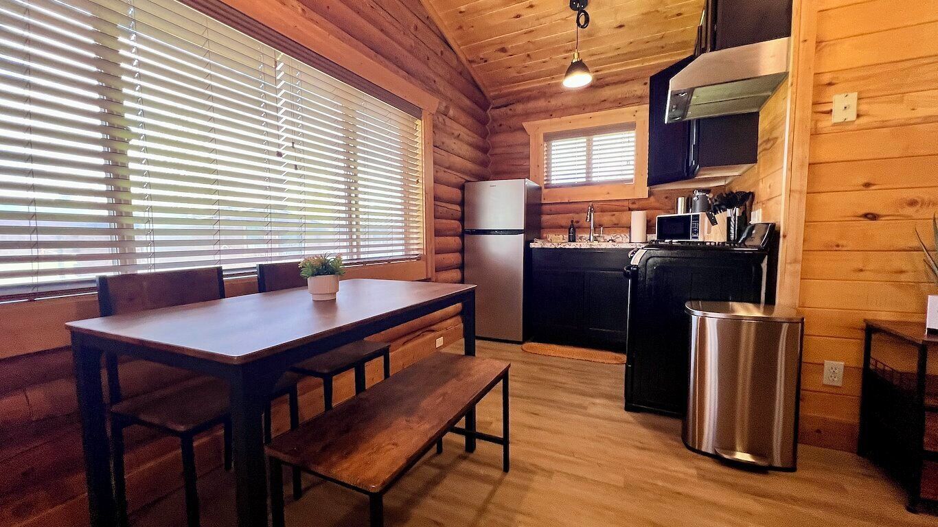 A wooden cabin interior with a dining table, benches, a kitchen area with appliances, and a trash can.