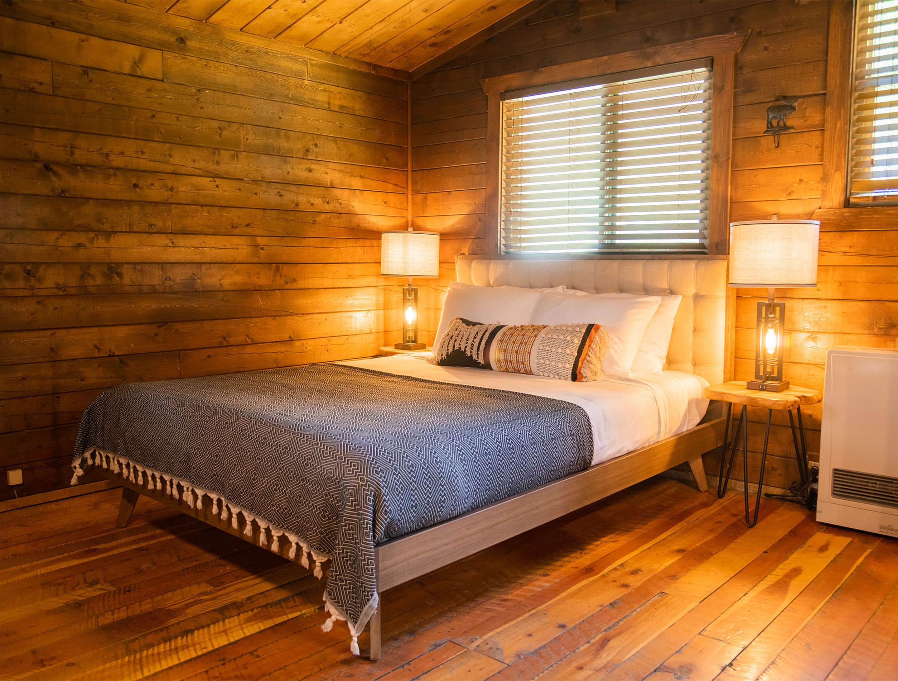 A wooden cabin bedroom with a bed, two bedside lamps, and a window with blinds.
