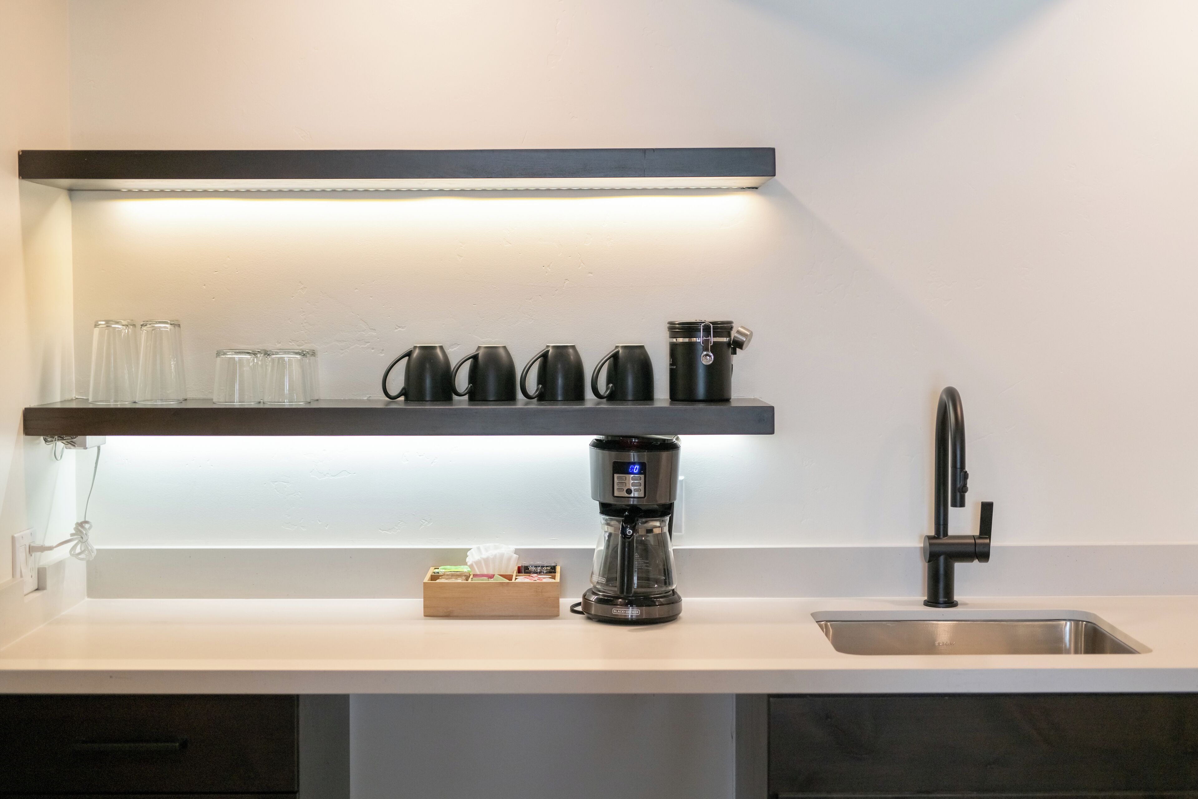 A modern kitchen with a coffee maker, a sink, and a shelf with glassware and coffee cups.