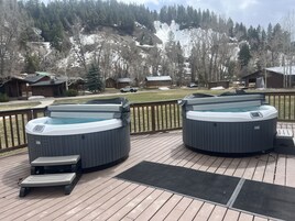 Dos jacuzzis en una terraza de madera con vista a una montaña nevada y casas al fondo.