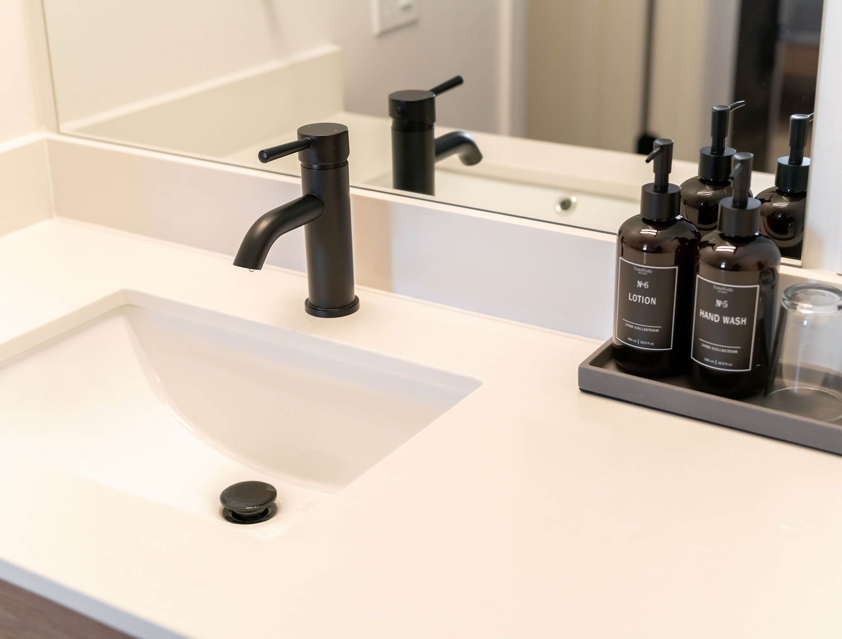 Un lavabo in bagno con un rubinetto moderno, uno specchio e un vassoio con flaconi di sapone liquido.