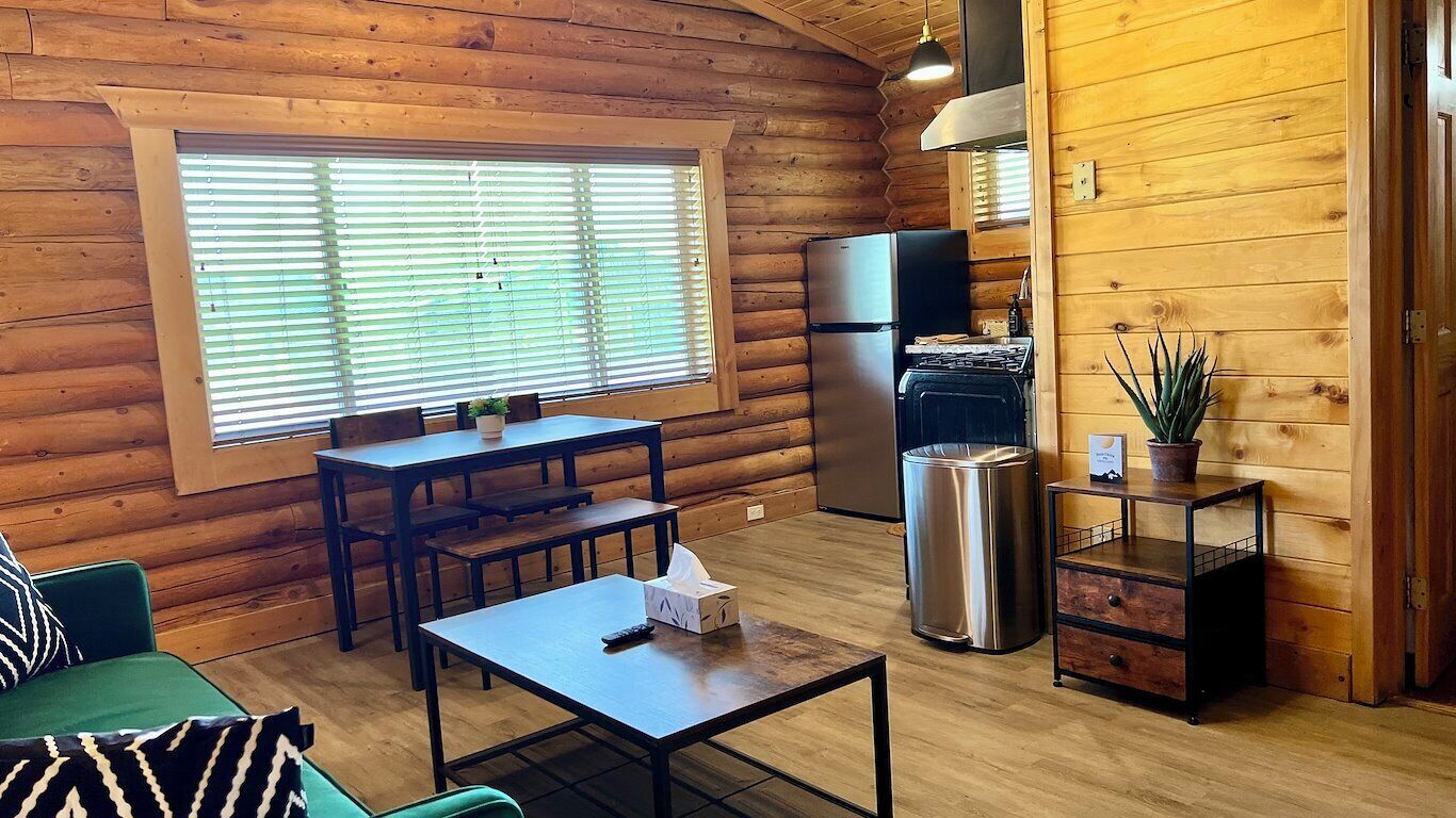Accogliente interno di una baita in legno con zona pranzo, cucina completa di piano cottura e frigorifero, e finestra con persiane.