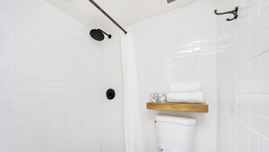 Een badkamer met witte betegelde wanden, een douche met een zwarte handdouche, een toilet met een wit zitje en een houten plankje met een handdoek erop.