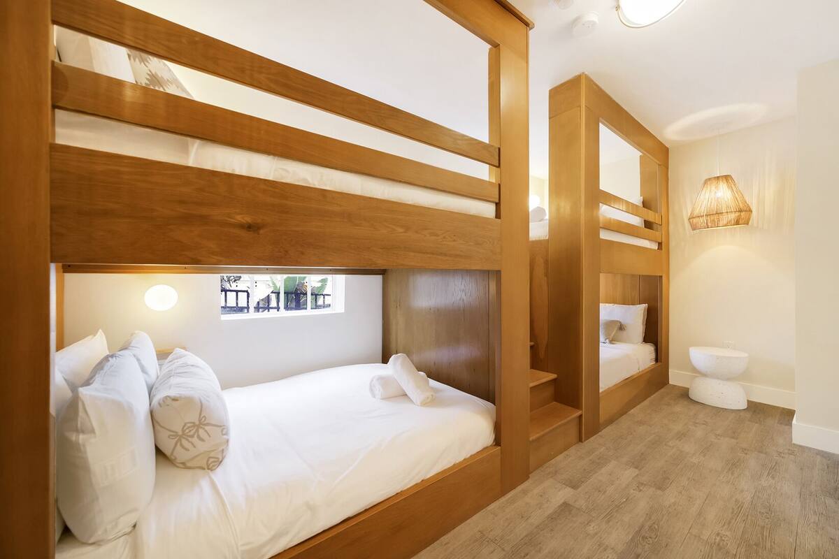 A bunk bed room with a wooden headboard, a window with a view, a bathroom with a toilet and sink, and a hanging light fixture.