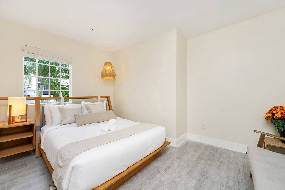 A bedroom with a large bed, a bedside table with a lamp, a window with a view of greenery, and a small table with flowers.