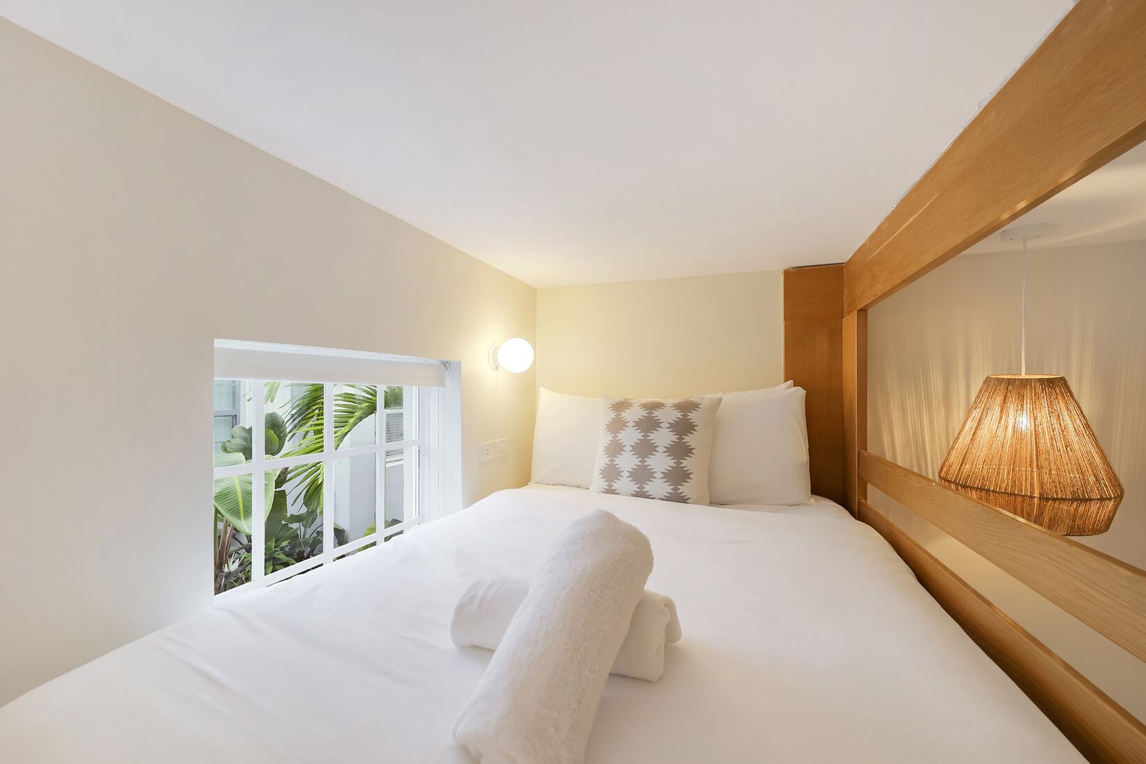 A hotel room with a bed, a window with a view of greenery, a wall-mounted light, and a hanging lamp.