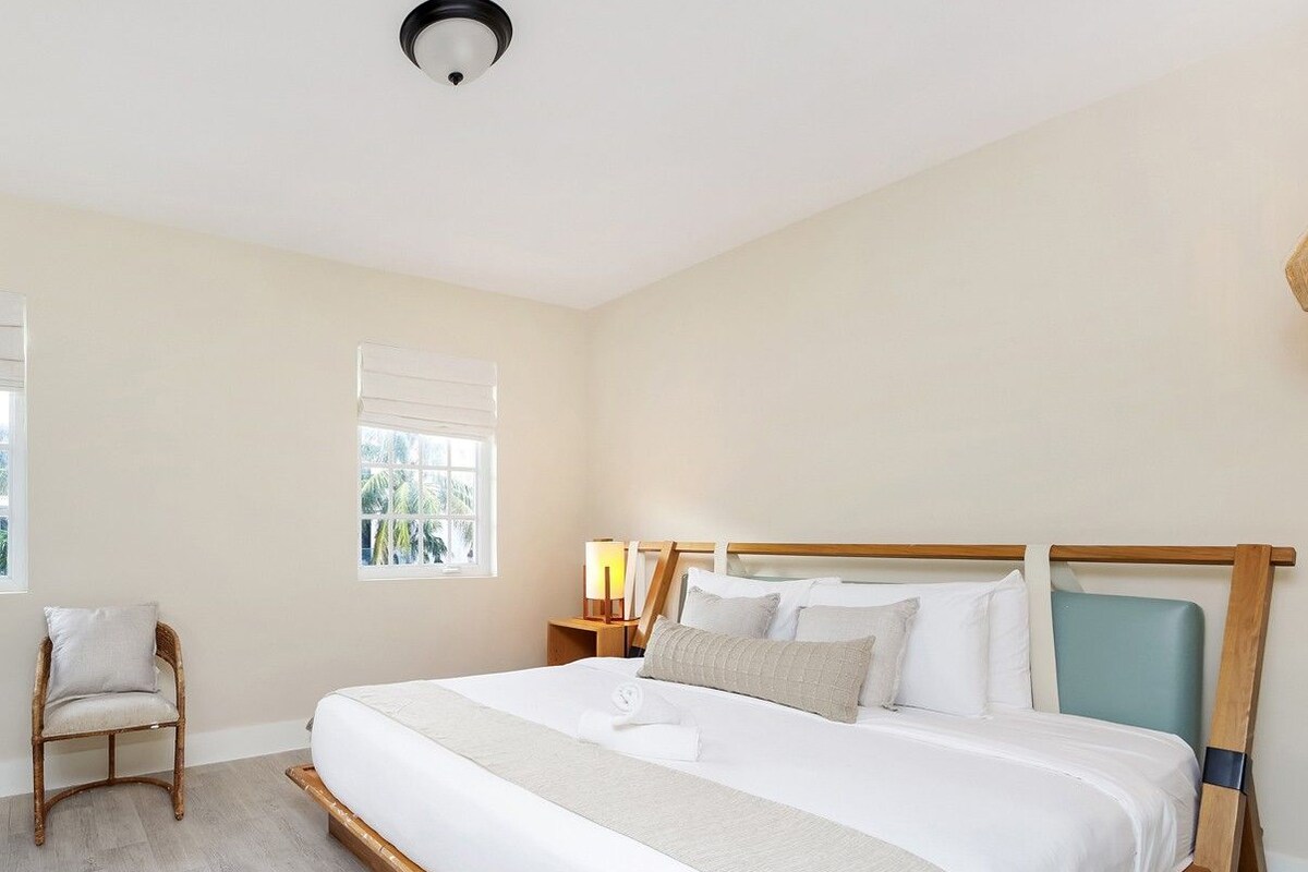 Quarto de hotel com uma cama grande, mesa de cabeceira com luminária, uma cadeira e janela com persianas.