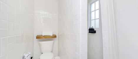 A bathroom with white tiled walls, a toilet, a shower area, and a window.