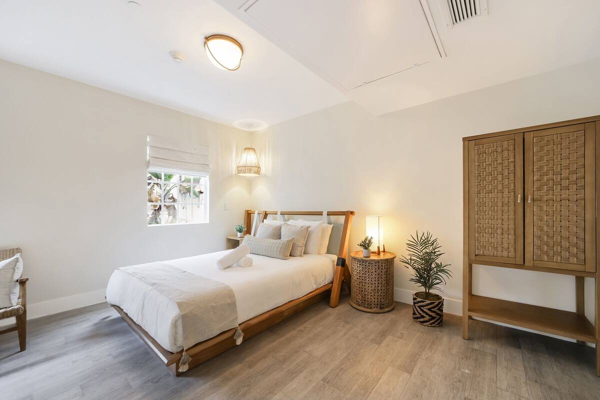 A bedroom with a wooden bed, a bedside lamp, a small round table, and a potted plant.