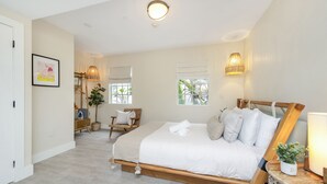 Een slaapkamer met een groot bed, een stoel, een nachtkastje met een lamp en een raam met uitzicht op groen.