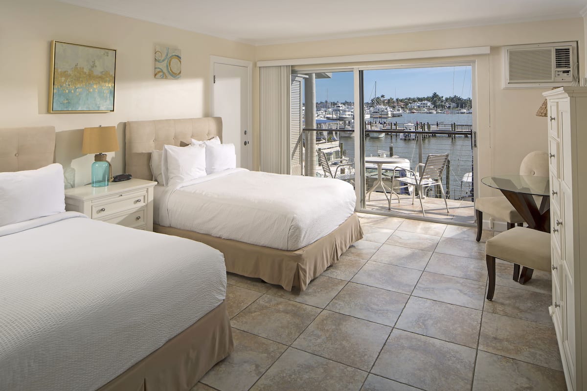 A hotel room with two beds, a balcony with a view of the marina, and a dining area.