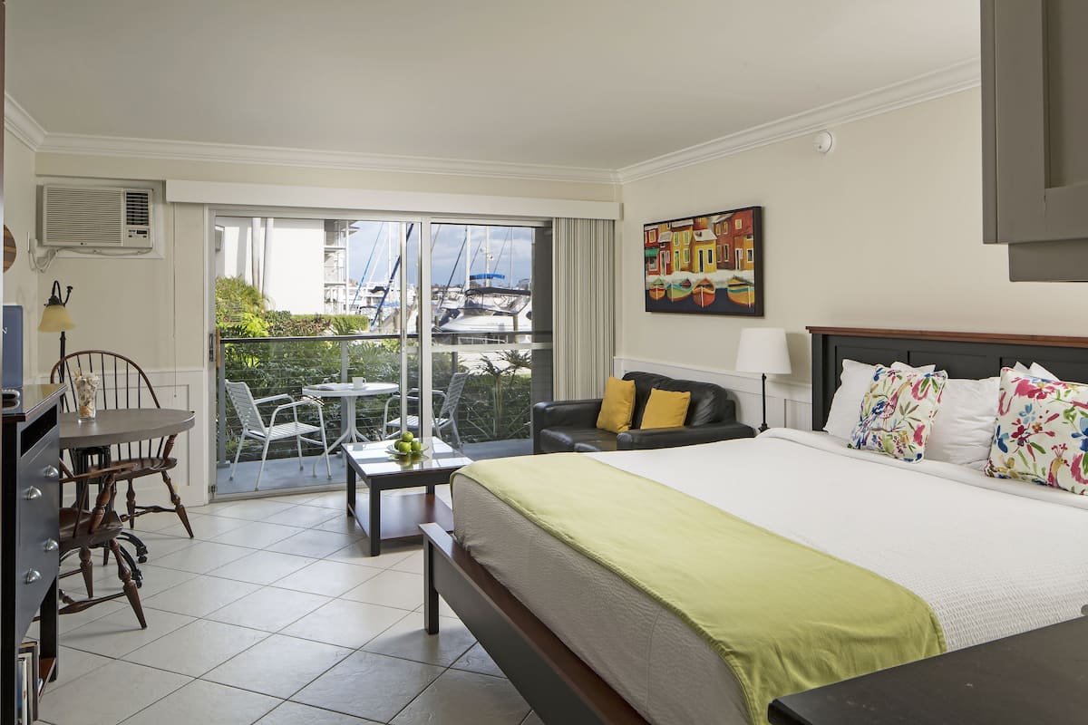 A hotel room with a large bed, a desk, a chair, a balcony with a view of boats, and a painting on the wall.