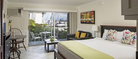 A hotel room with a large bed, a desk, a chair, a balcony with a view of boats, and a painting on the wall.