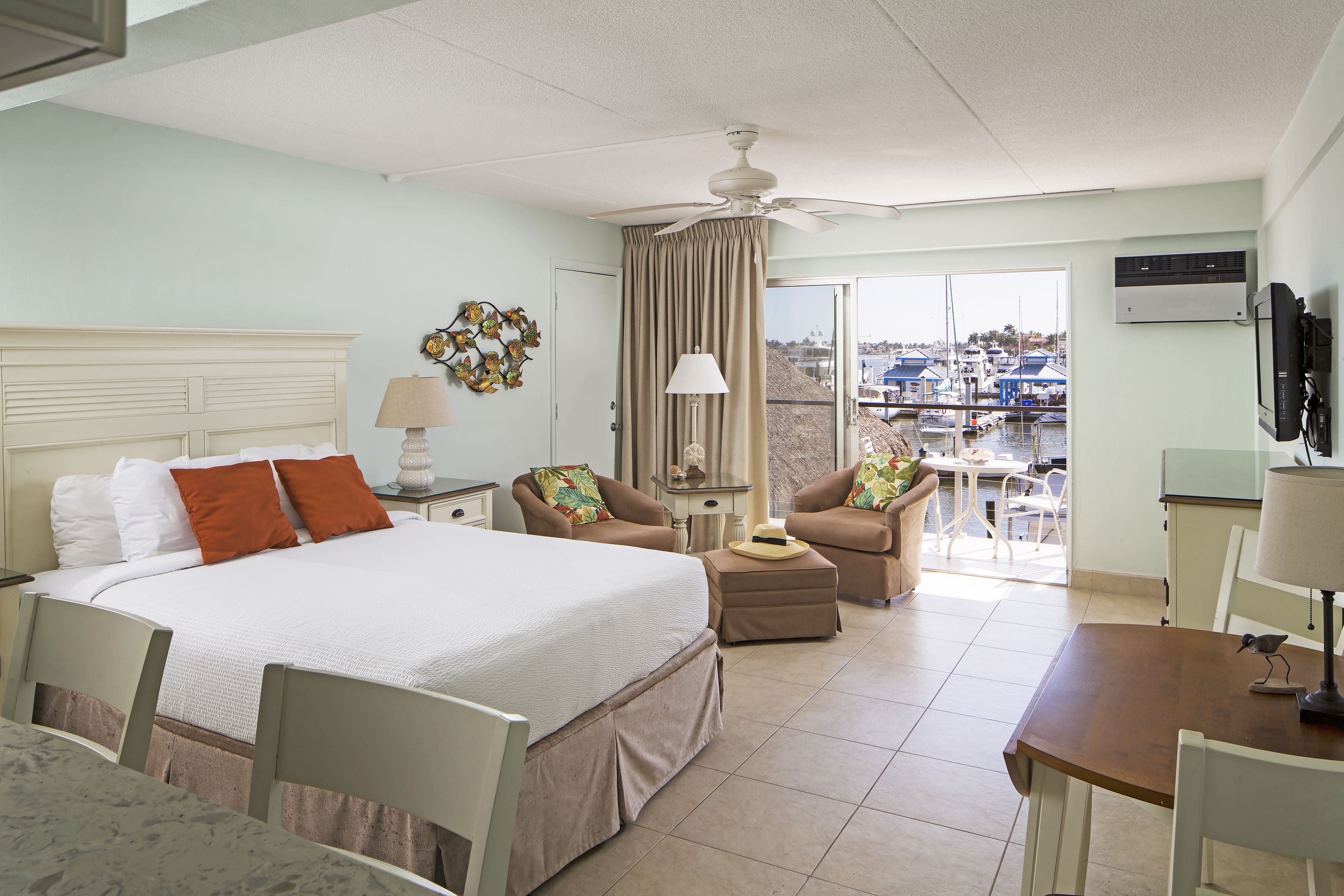 Habitación de hotel con cama, dos sillones, una mesita, ventilador de techo y vista al puerto deportivo a través de la ventana.