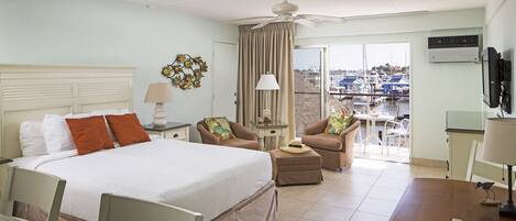 Habitación de hotel con cama, dos sillones, una mesita, ventilador de techo y vista al puerto deportivo a través de la ventana.
