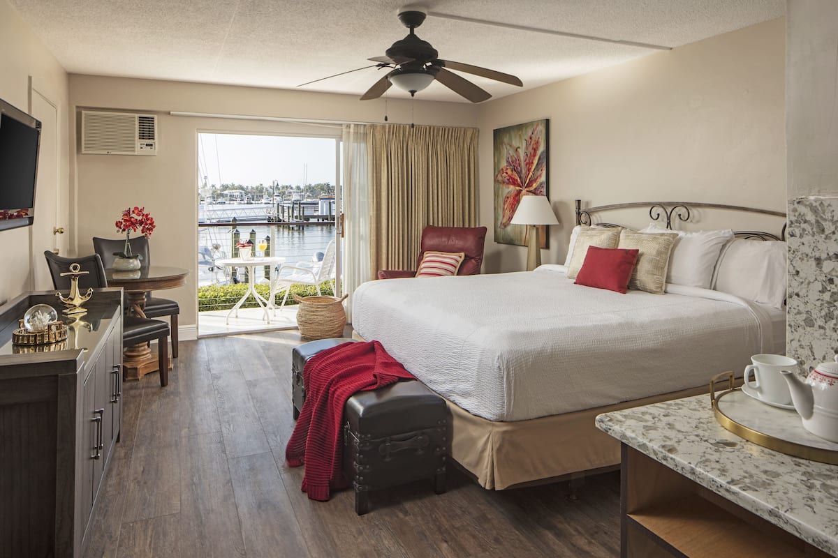 A hotel room with a bed, a desk, a chair, a small table, a ceiling fan, and a view of a marina.