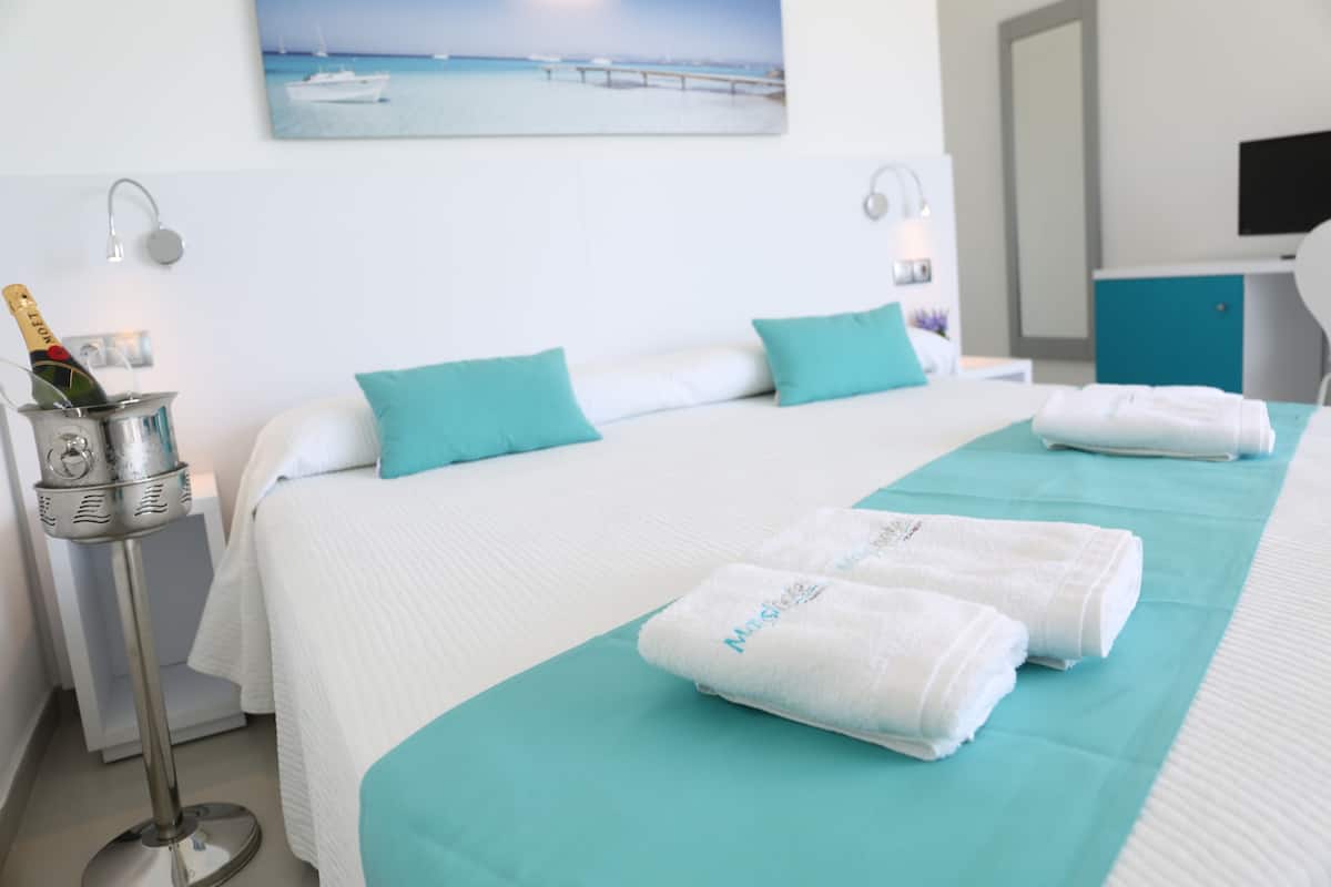 A hotel room with a bed, a minibar, a painting of a boat on the wall, and a television on a stand.