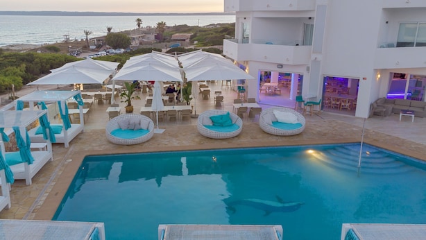A pool area with white lounge chairs and umbrellas, a clear view of the sea, and a building with balconies. - Hotel Maysi (Formentera)