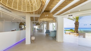 A modern hotel lobby with a reception desk, seating area, and a large window offering a view of the beach. - Hotel Maysi (Formentera)