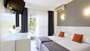 A hotel room with two beds, a TV, a desk, and a balcony with a view of greenery.