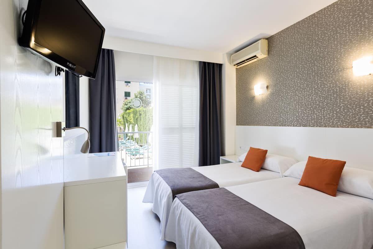 Ein Hotelzimmer mit zwei Betten, einem Fernseher, einem Schreibtisch und einem Balkon mit Blick auf Grünflächen.