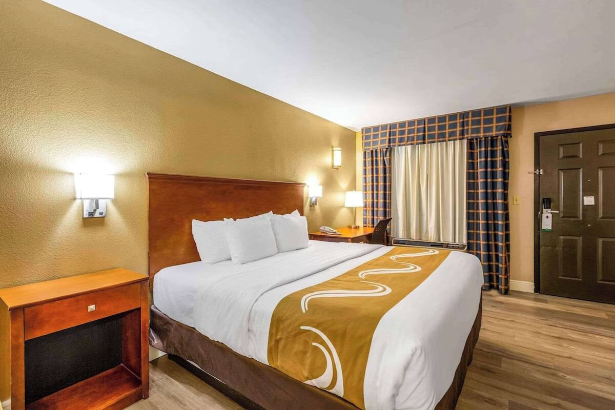 Quarto de hotel com uma cama grande, cabeceira de madeira, mesas de cabeceira e um criado-mudo.