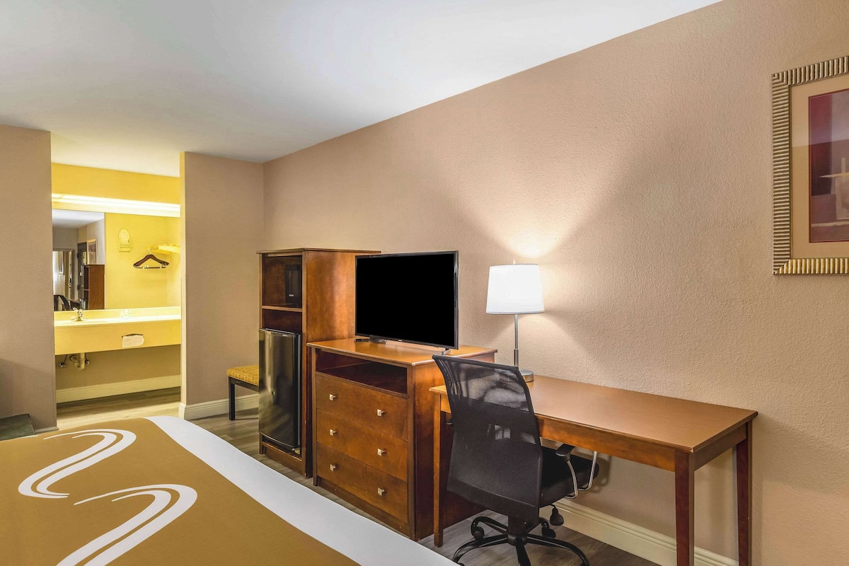 Quarto de hotel com escrivaninha de madeira, televisão, cadeira, abajur e guarda-roupa.