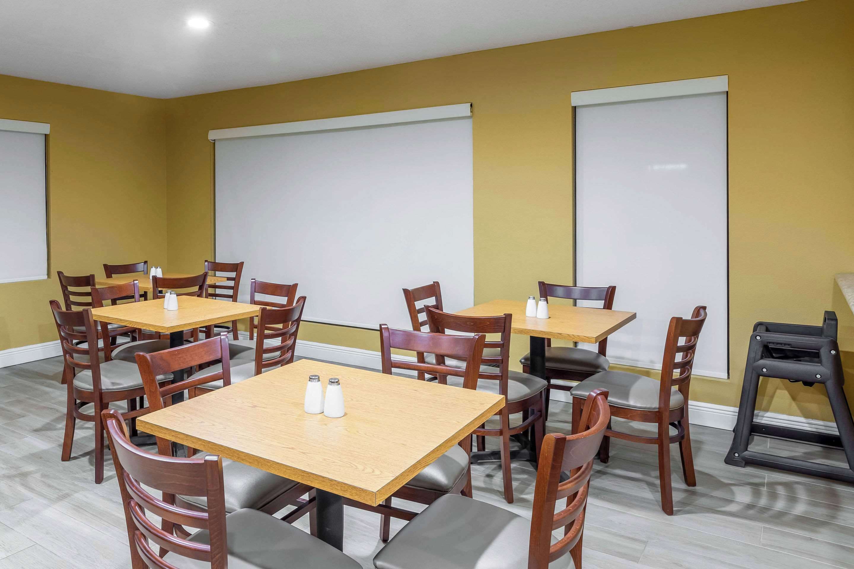 Un espace repas avec des tables et des chaises en bois, des salières blanches sur chaque table et de grandes fenêtres équipées de stores.